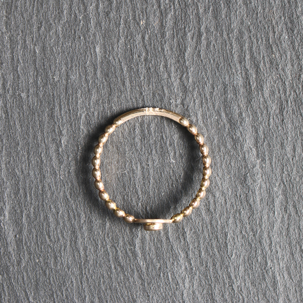 A thin gold beaded ring with a small central connector, displayed on a textured gray slate surface.