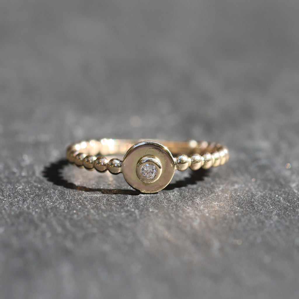 A gold ring with a textured beaded band and a central round gold disc featuring a small clear gemstone, resting on a dark gray textured surface.