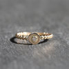 A gold ring with a textured beaded band and a central round gold disc featuring a small clear gemstone, resting on a dark gray textured surface.