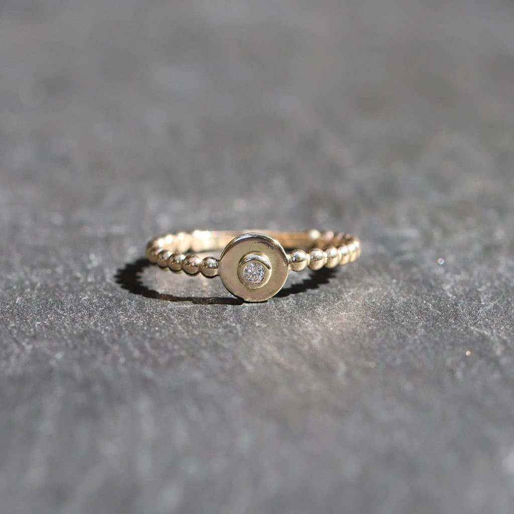 A delicate gold ring with a beaded band, a central flat circular disc, and a small clear gemstone in the middle, resting on a textured gray surface.