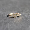 A delicate gold ring with a beaded band, a central flat circular disc, and a small clear gemstone in the middle, resting on a textured gray surface.