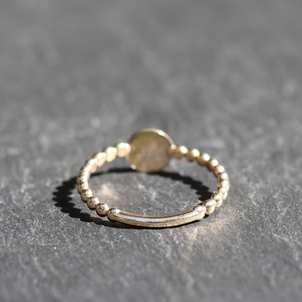 A delicate gold beaded ring with a smooth central band and a small round disc detail, resting on a textured gray slate surface.