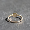 A delicate gold beaded ring with a smooth central band and a small round disc detail, resting on a textured gray slate surface.