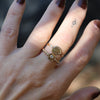 A close-up of a hand wearing two delicate gold rings. The primary ring features a small round clear gemstone, while the ring beneath it has a textured, organic gold shape with a tiny central stone.
